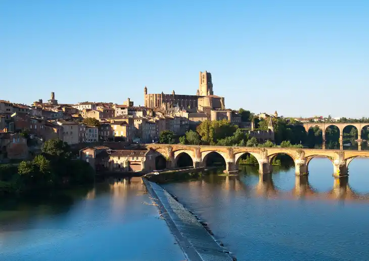 Paysage albigeois montrant une chaussée sur la rivière Tarn au premier plan, surplombée par les bâtiments du Palais de la Berbie et le clocher de la cathédrale sous un ciel ensoleillé.