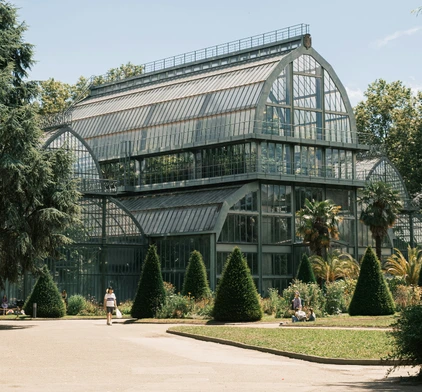 Les grandes serres historiques en fer forgé et verre du Jardin de la Tête d'Or, entourées de jardins paysagers et de conifères.