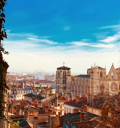 Vue sur les toits de tuiles rouges du Vieux Lyon avec la Cathédrale Saint-Jean au premier plan et la Grande Roue de la Place Bellecour visible au loin sous un ciel dégradé.