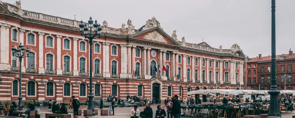 The facade of Toulouse's Capitole building, with its red bricks and columns, overlooking the lively square with its market stalls.