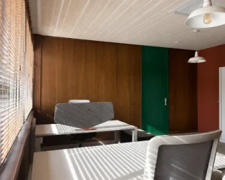 Bureau privatif lumineux avec revêtement mural en bois, porte verte, et bureau blanc équipé d'un écran acoustique près d'une fenêtre à stores vénitiens.