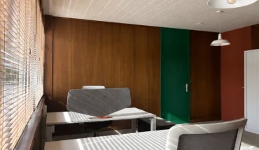 Bureau privatif lumineux avec revêtement mural en bois, porte verte, et bureau blanc équipé d'un écran acoustique près d'une fenêtre à stores vénitiens.