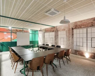 Vue de biais d'une salle de réunion aménagée en U montrant des chaises en bois design, des murs en briques et de larges baies vitrées à encadrement vert.