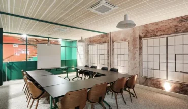 Vue de biais d'une salle de réunion aménagée en U montrant des chaises en bois design, des murs en briques et de larges baies vitrées à encadrement vert.