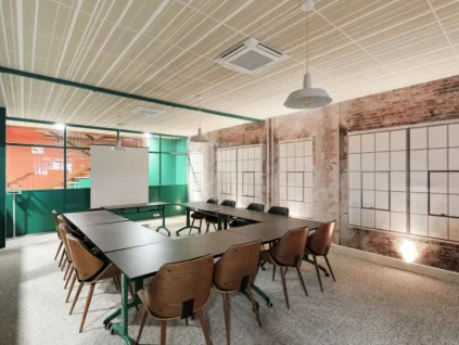 Vue de biais d'une salle de réunion aménagée en U montrant des chaises en bois design, des murs en briques et de larges baies vitrées à encadrement vert.