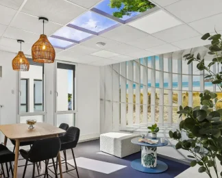 Espace détente du centre BURO Club Vannes Ténénio, avec table haute en bois, chaises noires, suspensions en osier, pouf blanc et décor de plage derrière une cloison à lamelles.