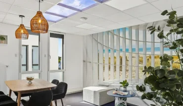 Espace détente du centre BURO Club Vannes Ténénio, avec table haute en bois, chaises noires, suspensions en osier, pouf blanc et décor de plage derrière une cloison à lamelles.