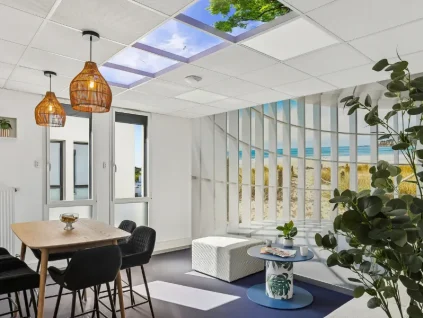 Espace détente du centre BURO Club Vannes Ténénio, avec table haute en bois, chaises noires, suspensions en osier, pouf blanc et décor de plage derrière une cloison à lamelles.