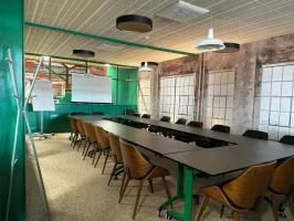 Salle de réunion de style industriel configurée en U, avec des tables noires, un mur en briques apparentes, une verrière verte et un écran de projection.