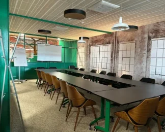 Salle de réunion de style industriel configurée en U, avec des tables noires, un mur en briques apparentes, une verrière verte et un écran de projection.