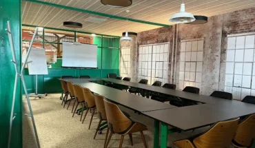 Salle de réunion de style industriel configurée en U, avec des tables noires, un mur en briques apparentes, une verrière verte et un écran de projection.