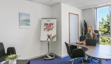 Bureau privatif du centre BURO Club Vannes Ténénio : une femme travaille sur ordinateur portable face à une fenêtre, à côté d'un paperboard et d'un coin salon.