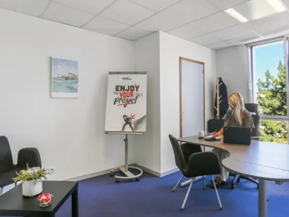Bureau privatif du centre BURO Club Vannes Ténénio : une femme travaille sur ordinateur portable face à une fenêtre, à côté d'un paperboard et d'un coin salon.
