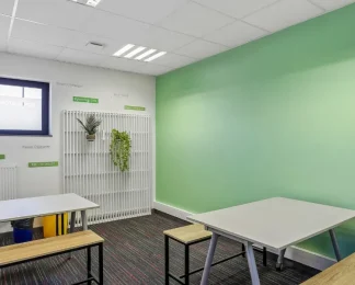 Espace de pause du centre BURO Club Vannes Laroiseau, avec tables blanches, bancs en bois, mur vert et plantes suspendues sur un treillis blanc.