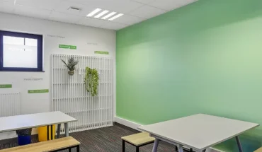 Espace de pause du centre BURO Club Vannes Laroiseau, avec tables blanches, bancs en bois, mur vert et plantes suspendues sur un treillis blanc.