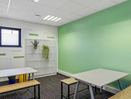 Espace de pause du centre BURO Club Vannes Laroiseau, avec tables blanches, bancs en bois, mur vert et plantes suspendues sur un treillis blanc.