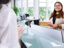 Réceptionniste souriante au comptoir d'accueil du Business Campus Mainz accueillant un client dans un espace bureau chaleureux et lumineux.