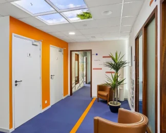 Couloir du centre BURO Club Vannes Ténénio avec moquette bleue à liseré orange, mur orange, fauteuils en cuir camel, plante verte et plafond imitant un ciel.