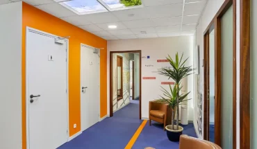 Couloir du centre BURO Club Vannes Ténénio avec moquette bleue à liseré orange, mur orange, fauteuils en cuir camel, plante verte et plafond imitant un ciel.