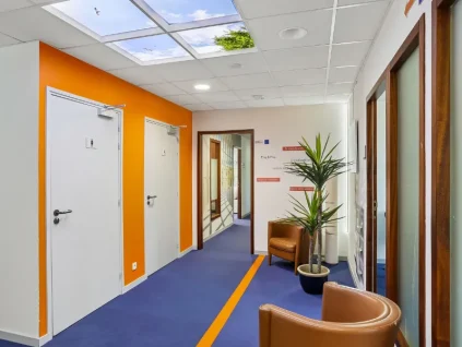 Couloir du centre BURO Club Vannes Ténénio avec moquette bleue à liseré orange, mur orange, fauteuils en cuir camel, plante verte et plafond imitant un ciel.