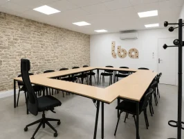 Salle de réunion en carré au BURO Club Partner Beaune avec tables en bois, chaises noires et mur en pierres apparentes.