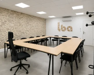 Salle de réunion en carré au BURO Club Partner Beaune avec tables en bois, chaises noires et mur en pierres apparentes.