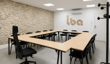 Salle de réunion en carré au BURO Club Partner Beaune avec tables en bois, chaises noires et mur en pierres apparentes.