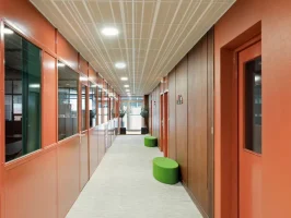 Couloir de circulation d'un centre d'affaires aux murs peints en terracotta, bordé par une verrière d'atelier verte et agrémenté de poufs d'assise verts.