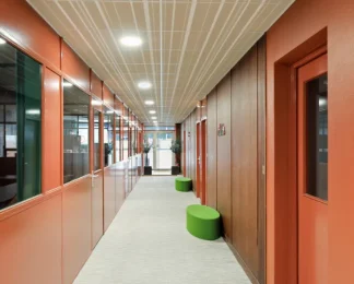 Couloir de circulation d'un centre d'affaires aux murs peints en terracotta, bordé par une verrière d'atelier verte et agrémenté de poufs d'assise verts.
