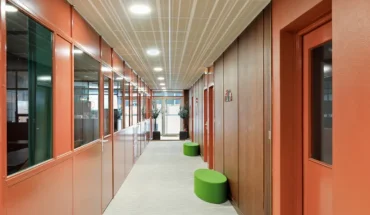 Couloir de circulation d'un centre d'affaires aux murs peints en terracotta, bordé par une verrière d'atelier verte et agrémenté de poufs d'assise verts.