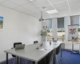 Salle de réunion du centre BURO Club Vannes Ténénio, avec tables grises, chaises noires, vidéoprojecteur, paperboard et fenêtres équipées de stores.