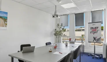 Salle de réunion du centre BURO Club Vannes Ténénio, avec tables grises, chaises noires, vidéoprojecteur, paperboard et fenêtres équipées de stores.