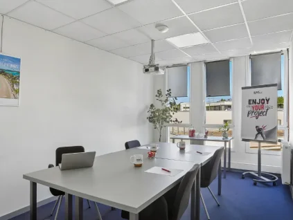 Salle de réunion du centre BURO Club Vannes Ténénio, avec tables grises, chaises noires, vidéoprojecteur, paperboard et fenêtres équipées de stores.
