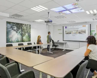 Salle de réunion du centre BURO Club Vannes Laroiseau, avec tables en U et vidéoprojecteur ; un intervenant présente un écran à deux participantes.