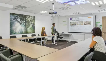 Salle de réunion du centre BURO Club Vannes Laroiseau, avec tables en U et vidéoprojecteur ; un intervenant présente un écran à deux participantes.