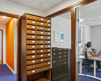 Casiers à courrier en bois du centre BURO Club Vannes Ténénio, à côté d'un bureau vitré où une femme travaille sur ordinateur.