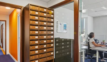 Casiers à courrier en bois du centre BURO Club Vannes Ténénio, à côté d'un bureau vitré où une femme travaille sur ordinateur.