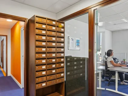 Casiers à courrier en bois du centre BURO Club Vannes Ténénio, à côté d'un bureau vitré où une femme travaille sur ordinateur.