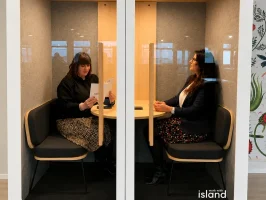 Deux professionnelles assises face à face discutant dans une cabine acoustique insonorisée vitrée (phone booth) au sein d'un espace de travail.