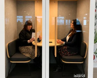 Deux professionnelles assises face à face discutant dans une cabine acoustique insonorisée vitrée (phone booth) au sein d'un espace de travail.