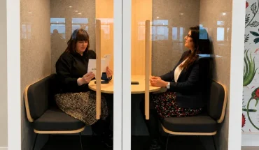 Deux professionnelles assises face à face discutant dans une cabine acoustique insonorisée vitrée (phone booth) au sein d'un espace de travail.