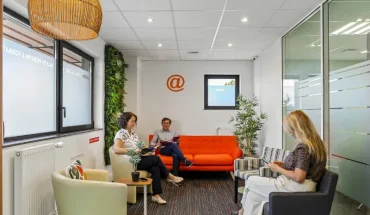 Salon d'accueil du centre BURO Club Vannes Laroiseau, avec canapé orange, fauteuils, mur végétal et suspension en osier ; trois personnes patientent en lisant.