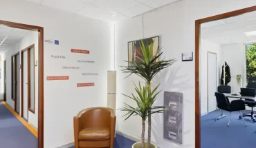 Hall du centre BURO Club Vannes Ténénio avec signalétique des services (bureaux équipés, salles de réunion et de formation, coworking), fauteuil camel et plante, ouvert sur un bureau.