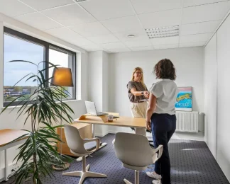 Bureau privatif lumineux du centre BURO Club Vannes Laroiseau, avec bureau en bois, deux chaises blanches et grande fenêtre ; deux femmes échangent debout.