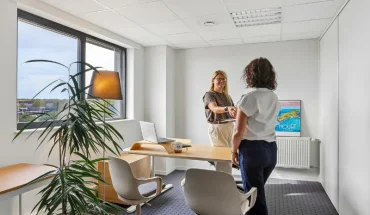 Bureau privatif lumineux du centre BURO Club Vannes Laroiseau, avec bureau en bois, deux chaises blanches et grande fenêtre ; deux femmes échangent debout.