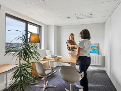 Bureau privatif lumineux du centre BURO Club Vannes Laroiseau, avec bureau en bois, deux chaises blanches et grande fenêtre ; deux femmes échangent debout.