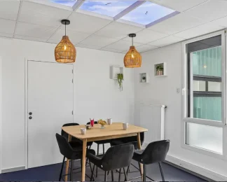 Coin repas du centre BURO Club Vannes Ténénio, avec table en bois, chaises noires, suspensions en osier, étagères avec plantes et plafond imitant un ciel.