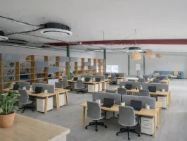 Espace de coworking en open space aménagé avec des rangées de bureaux en bois, des séparateurs acoustiques gris et une grande étagère de séparation, sous un plafond de style industriel.