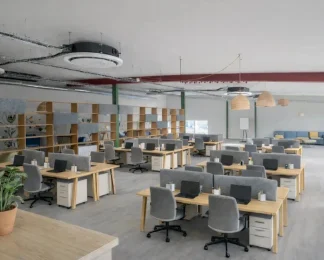 Espace de coworking en open space aménagé avec des rangées de bureaux en bois, des séparateurs acoustiques gris et une grande étagère de séparation, sous un plafond de style industriel.
