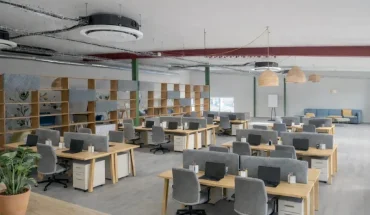 Espace de coworking en open space aménagé avec des rangées de bureaux en bois, des séparateurs acoustiques gris et une grande étagère de séparation, sous un plafond de style industriel.